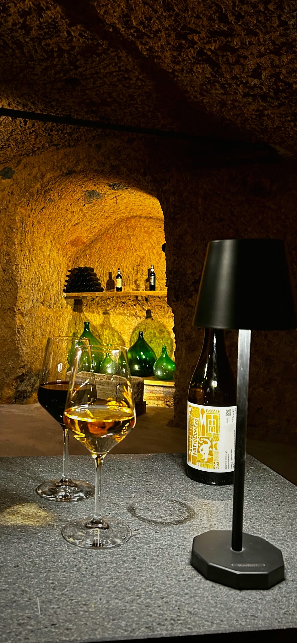 Two glasses of wine on a stone table in the glowing tuff cellar, antique demijohns resting in the niche behind.